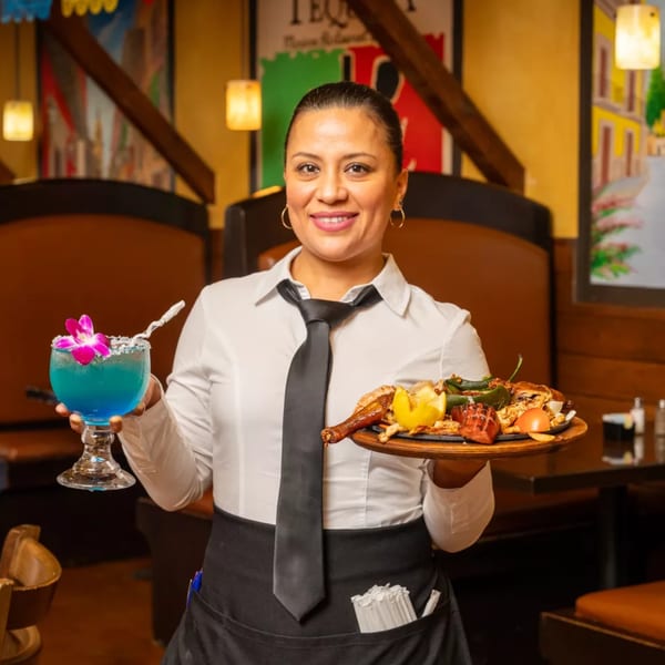 A smiling Grand Tequila server holds a blue margarita and a sizzling fajita platter.