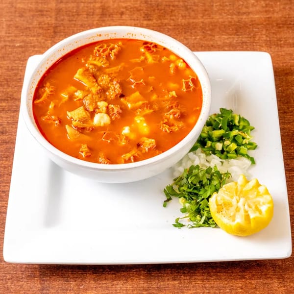 Menudo soup in a white bowl with a side of chopped onions, cilantro, jalapeños, and a lemon.