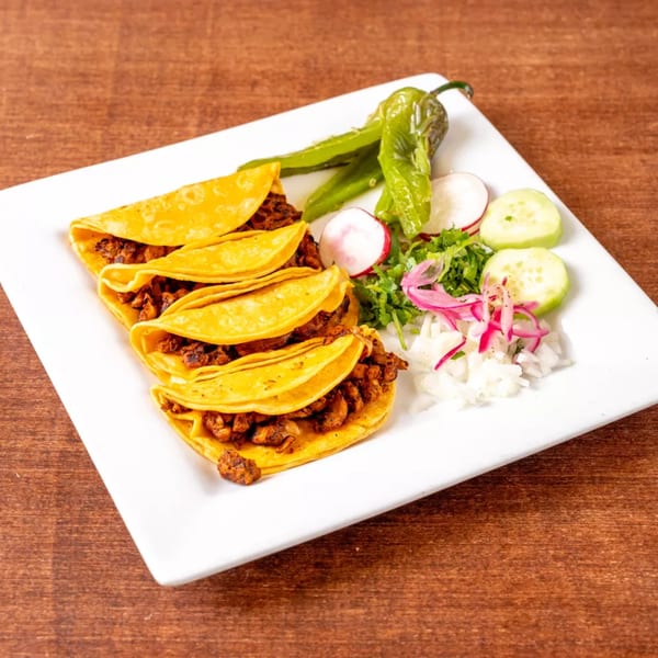 Four tacos with meat filling, green peppers, radishes, cucumbers, and onions on a white plate.