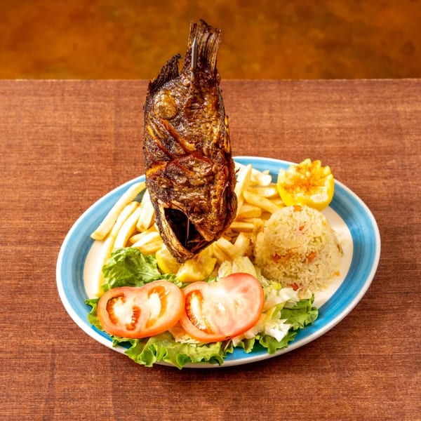 A whole fried fish stands upright on a plate with fries, rice, salad, and lemon.