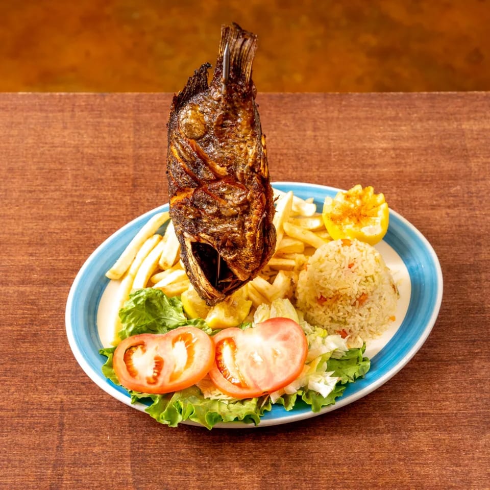 A whole fried fish stands upright on a plate with fries, rice, salad, and lemon.