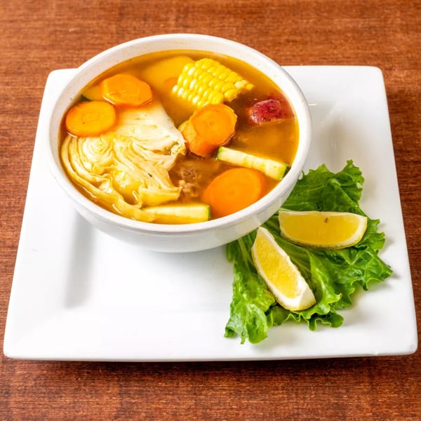 Hearty Mexican soup with corn, carrots, cabbage, and zucchini, garnished with lettuce and lemon wedges.