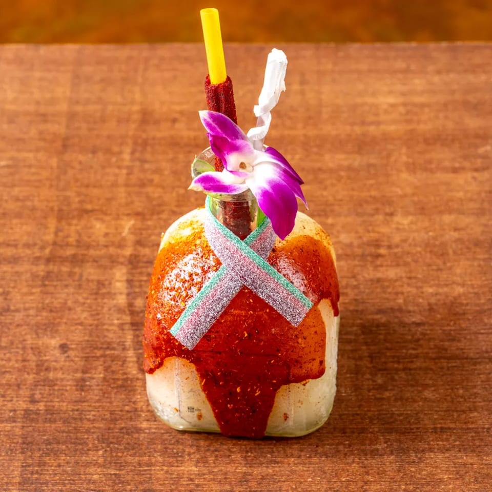 A bottle drink with red chili powder, gummy candy, a yellow straw, and a purple orchid.