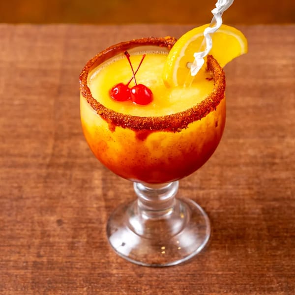 A vibrant yellow and orange drink in a stemmed glass, garnished with cherries, an orange slice, and a straw.